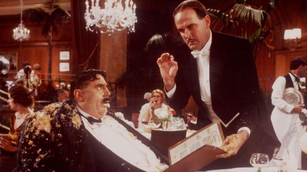 Terry Jones as Mr Creosote (left) and John Cleese standing, stained with vomit, holding a menu in front of him.