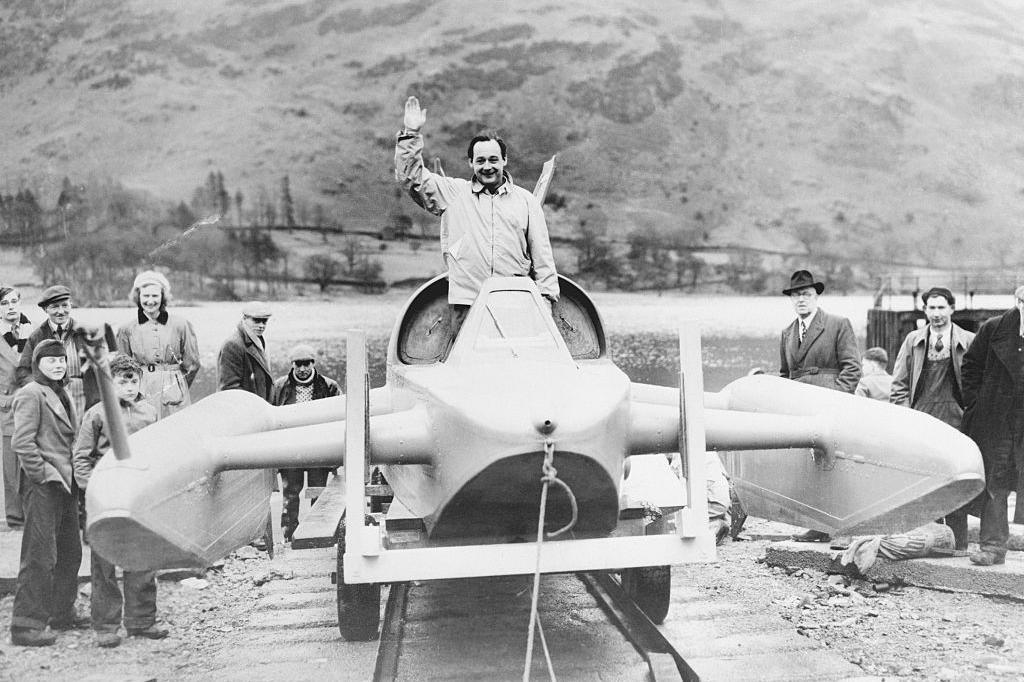 Black-and-white photograph of Donald Campbell waving from the cockpit of Bluebird at the shores of Ullswater in 1955. The lake can be seen in the background and several members of the public are standing next to the craft.