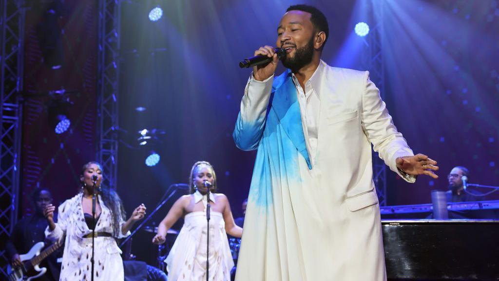 John Legend performs on stage in Kigali with instrumentalists and two backing singers. Legend wears a white and blue tunic - Friday 21 February 2025.