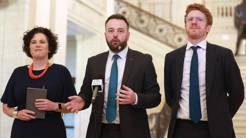 Three people standing in front of microphone. A woman on the left with short brown curly hair wearing a black dress an an orange necklace. A brown haired man wearing a suit with a teal tie in the middle and a ginger man wearing a black suit with a navy tie on the right.