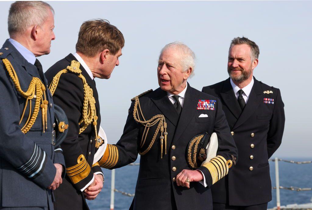 The King in black military uniform and holding a cap standing with three other men also in military uniform.