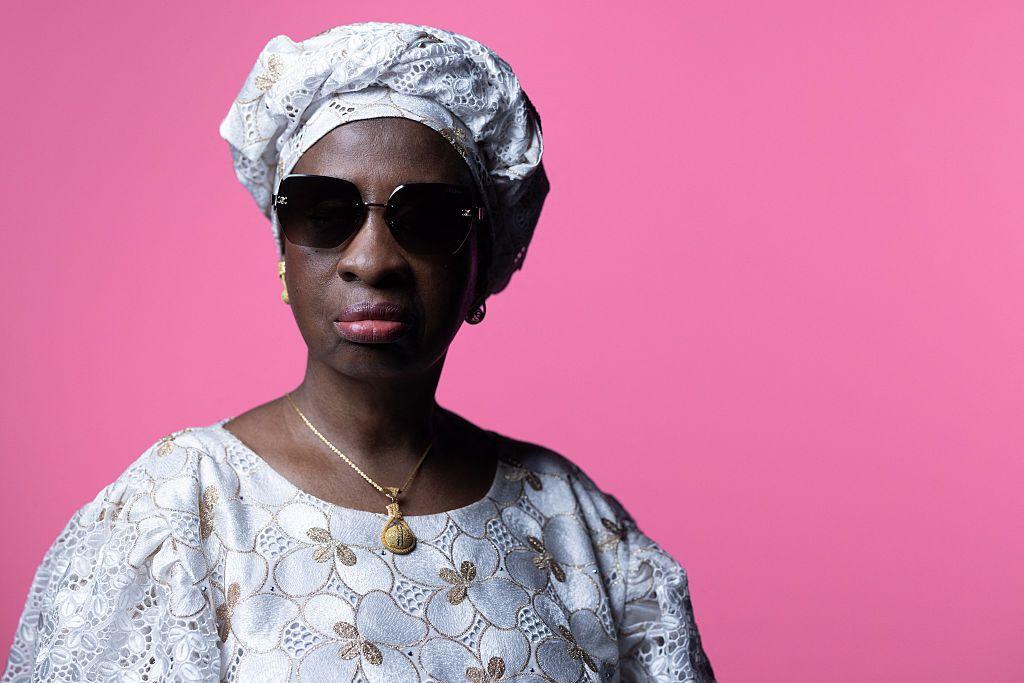 A woman wearing a matching lace dress and headwrap stands in front of a pink background.