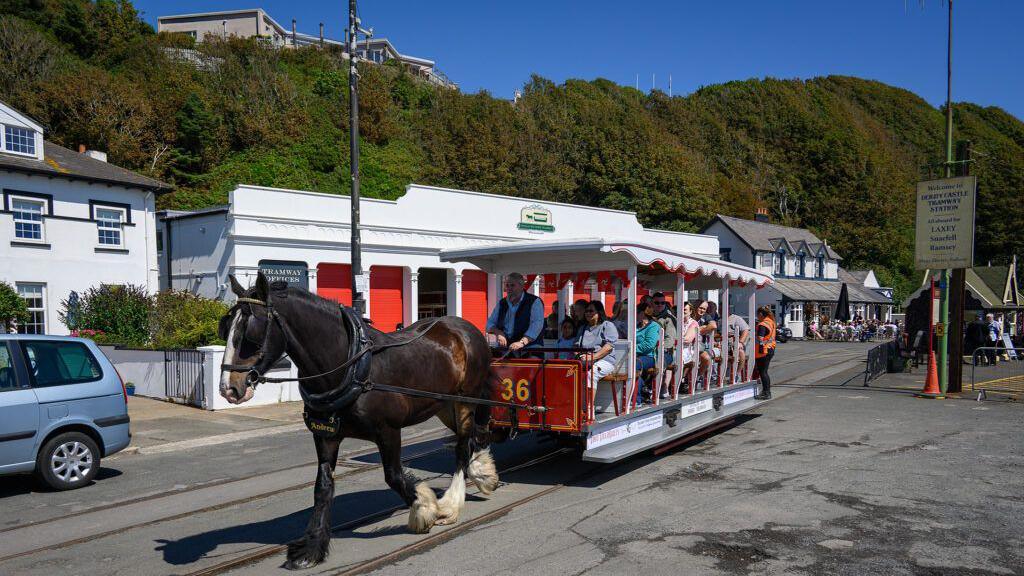 A brown horse pulls a Victorian tram car full of passengers as it leaves Derby Castle, with the tram storage depot in the background.