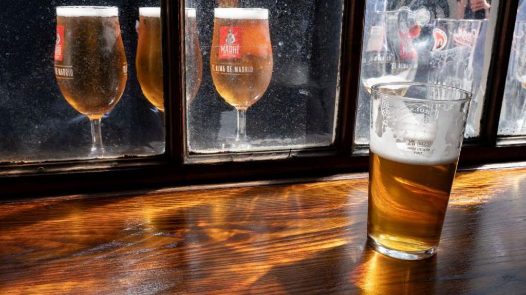 A half-drank pint of lager sits on a bar, in the background there are three full pints