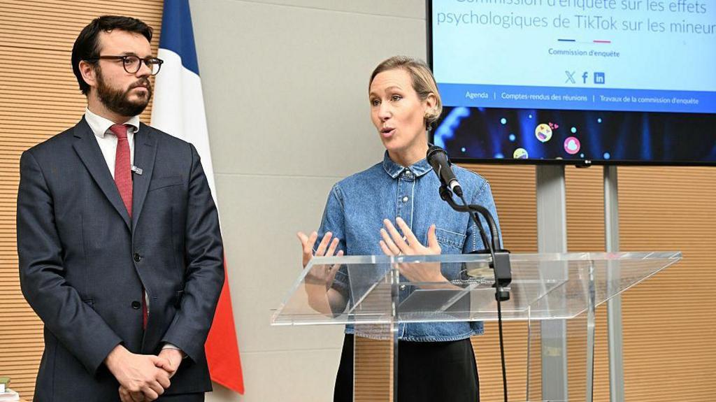 A woman in a denim shirt speaks at a lectern on the right while a man in a suit stands listening on her left