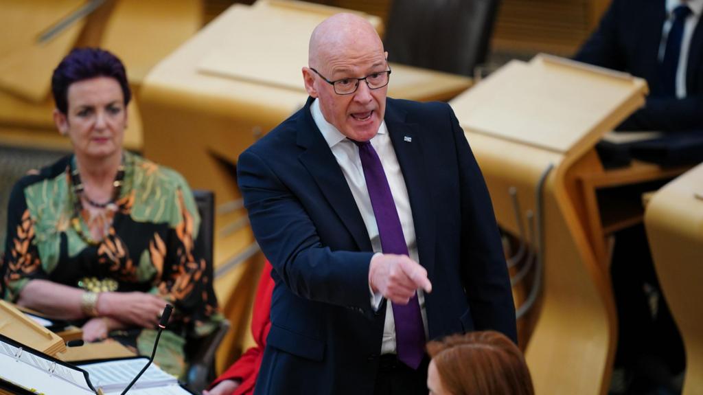 John Swinney points angrily in the chamber with Health Secretary Angela Constance sitting in the background
