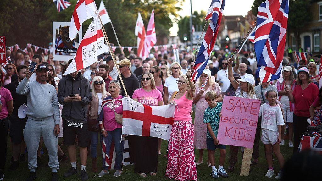 Large crowd of people waving Union Jack and St George flags outside council offices in Epping on 8 August 2025
