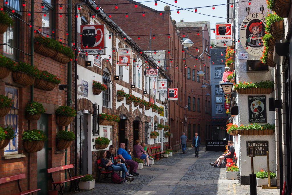 Exterior of a red brick and white bar with numerous vintage alcohol adverts and hanging plants, there are people sat on red benches outside the venue, which is situated on a narrow cobbled street with red festoon lighting linking the buildings.