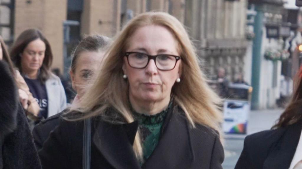 Kelly, wearing glasses and a black coat, walking up a street surrounded by other women in smart clothing