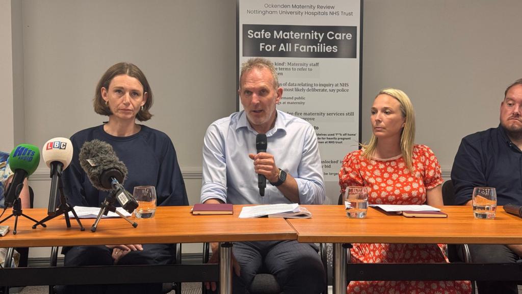 Two women and one man sit behind a long table, the man holds a microphone.