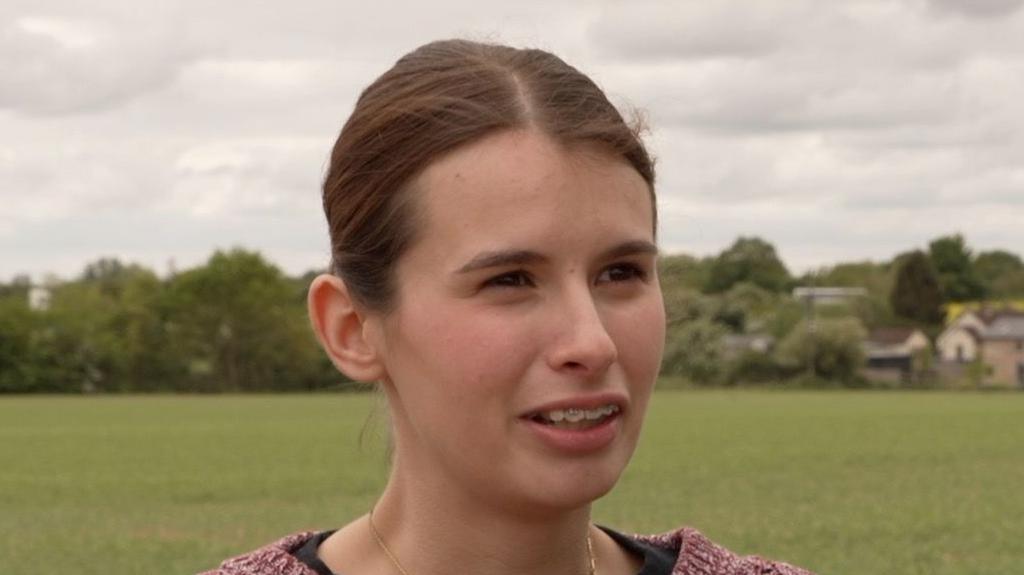 Adriana Hudson looks away from the camera while standing outside near a field. She has long dark hair that is tied back behind her head. She wears a red knitted jumper and black top underneath.