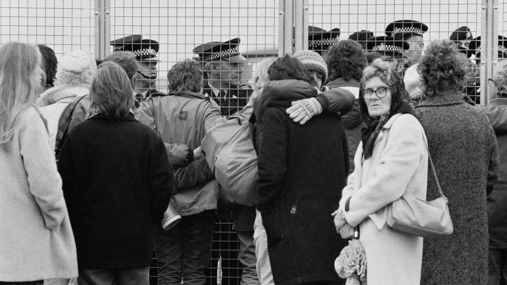Black and white archive photo of a group of women standing at the gates of RAF Greenham Common. Most have their backs to the camera, but one woman, who's wearing a headscarf and carrying a handbag, is looking towards the camera. Beyond the fence, there are about eight police officers, identifiable by their peaked caps. 