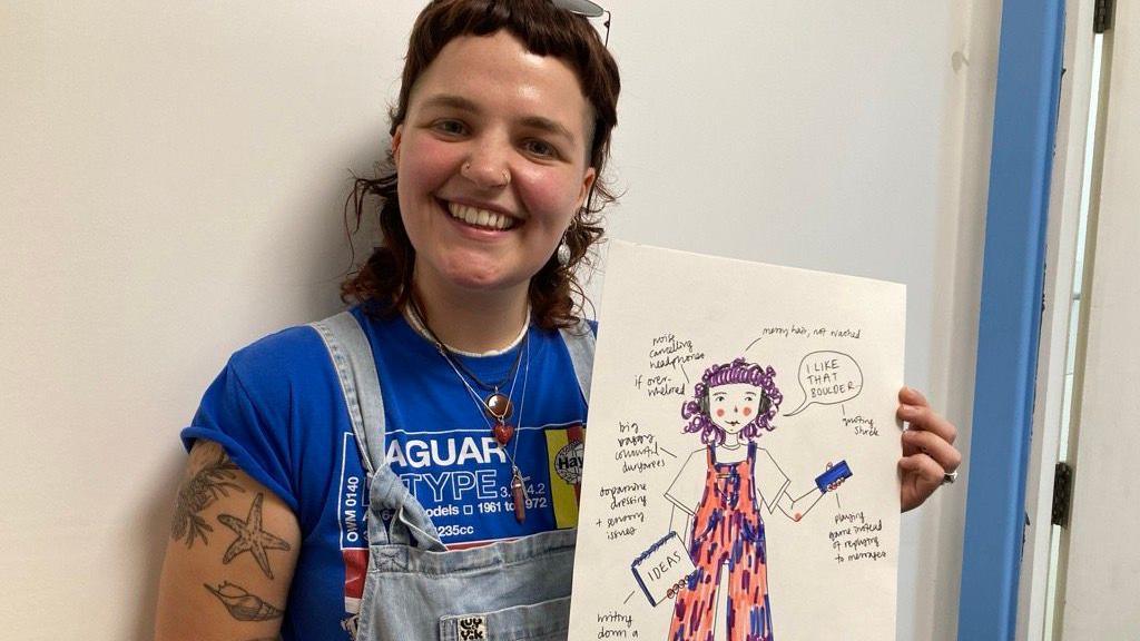 Bethany wearing a blue top and dungarees, holding the picture she drew of herself. 