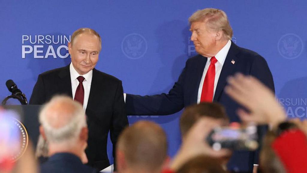 Vladimir Putin (L) walks on stage as Donald Trump (R) pats him on the back. In the blurred foreground are journalists raising their hands or holding up cameras