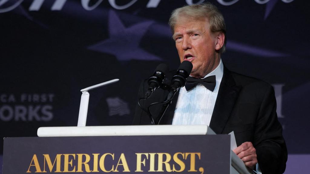 Donald Trump speaks in a tuxedo at a podium which has the words 'America first' on the front