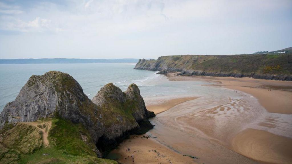 A look down on a large sandy beach. The tide is quite far in and water swells onto the golden sand. On either side are tall rocky cliffs.