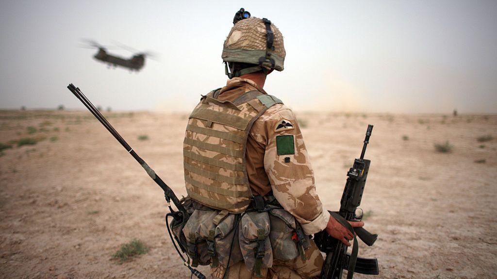 A British Army soldier from the 3rd Battalion, The Parachute Regiment, secures a helicopter landing strip in Maywand District in Kandahar Province, Afghanistan in 2008. The soldier is kneeling on the ground holding a rifle in his right hand, with his back to the camera. A military plane flies in the sky above him.