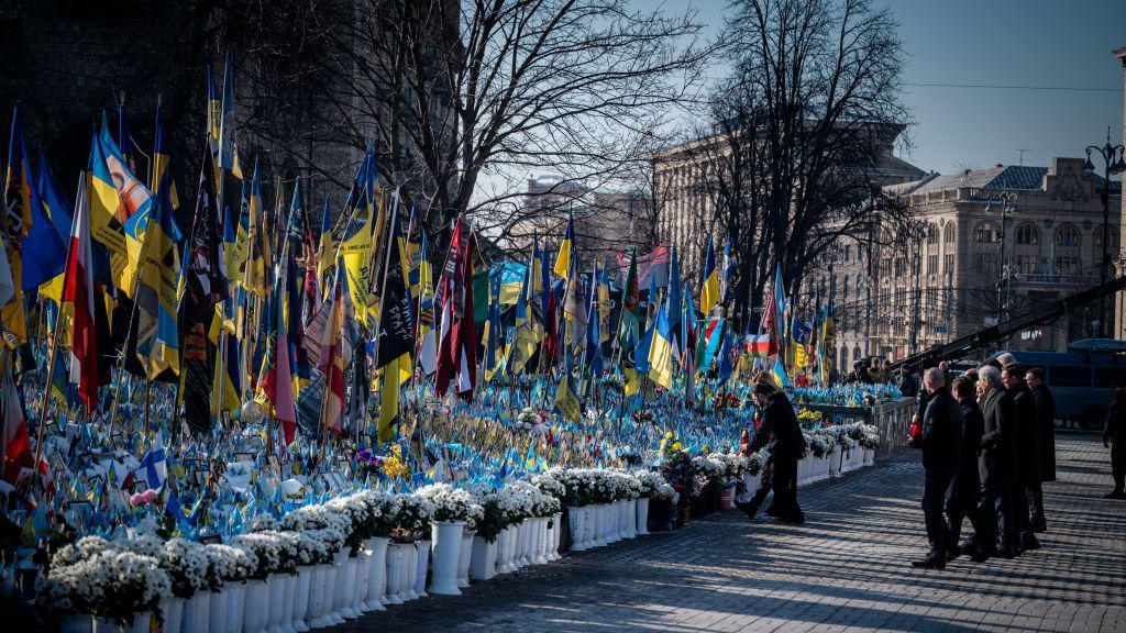 A group of people dressed in black watch as a man lays flowers by a collection of flowers and Ukrainian flags. They appear to be in a town square with buildings behind them. It's a sunny day and the sky is blue.