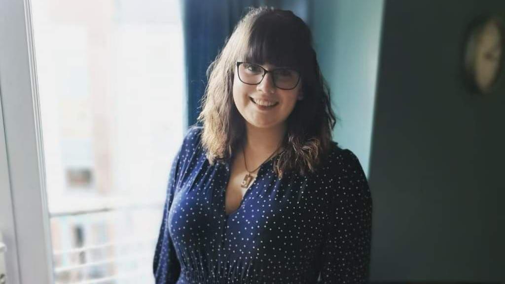 Rebecca is smiling and standing near glass doors. There is a clock on a wall behind her.