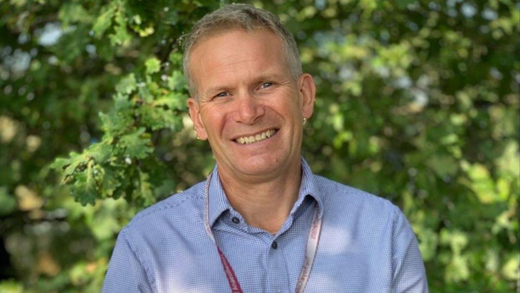 A publicity image of Dr Andrew Gregg wearing a pale blue shirt and a lanyard