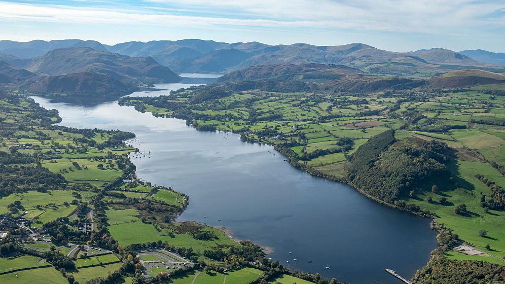 Aerial image of Ullswater. The large expanse of water is surrounded by green fields and hills.