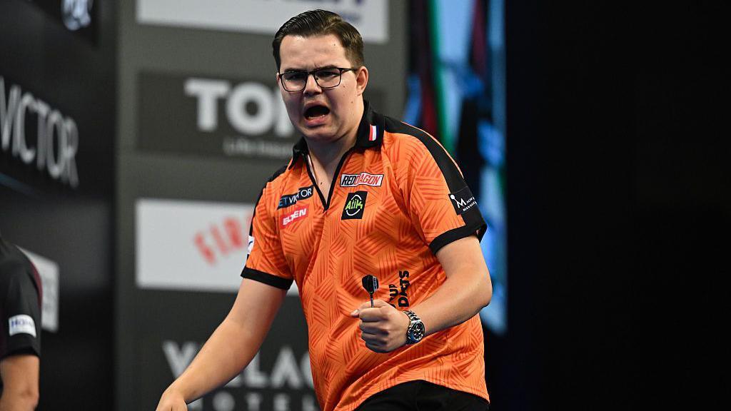 A man in glasses and an orange top shouting while holding darts