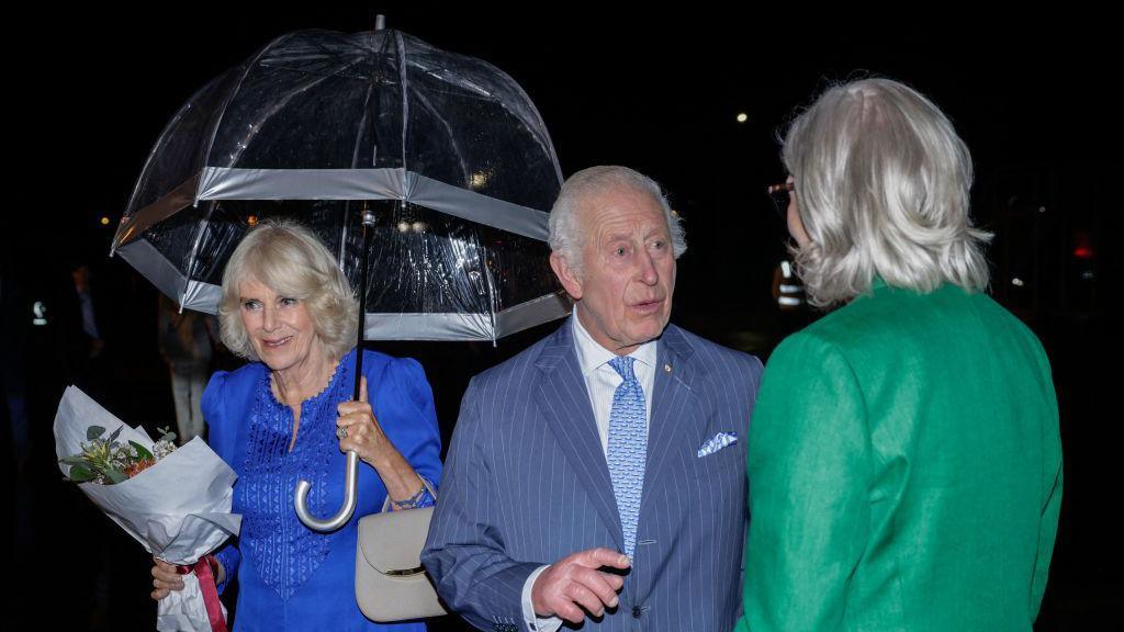 King Charles III speaks to Australia's Governor-General Sam Mostyn while Queen Camilla can be seen smiling behind him as she holds a posy of flowers 