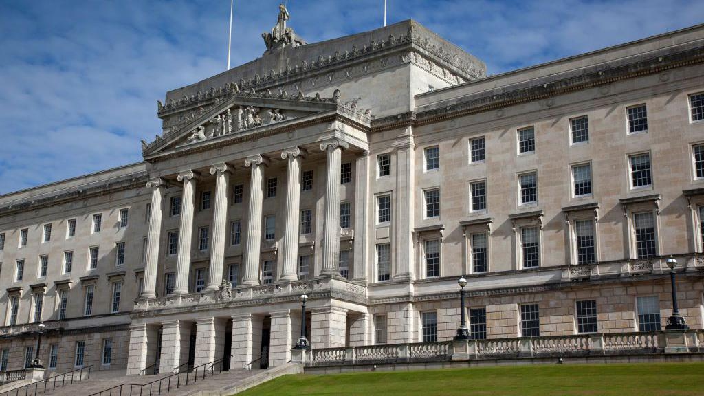 Stormont buildings from the outside. The sky in the background is bright and it looks like a sunny day. The building is cream stone with lots of windows and there are steps up to the entrance.