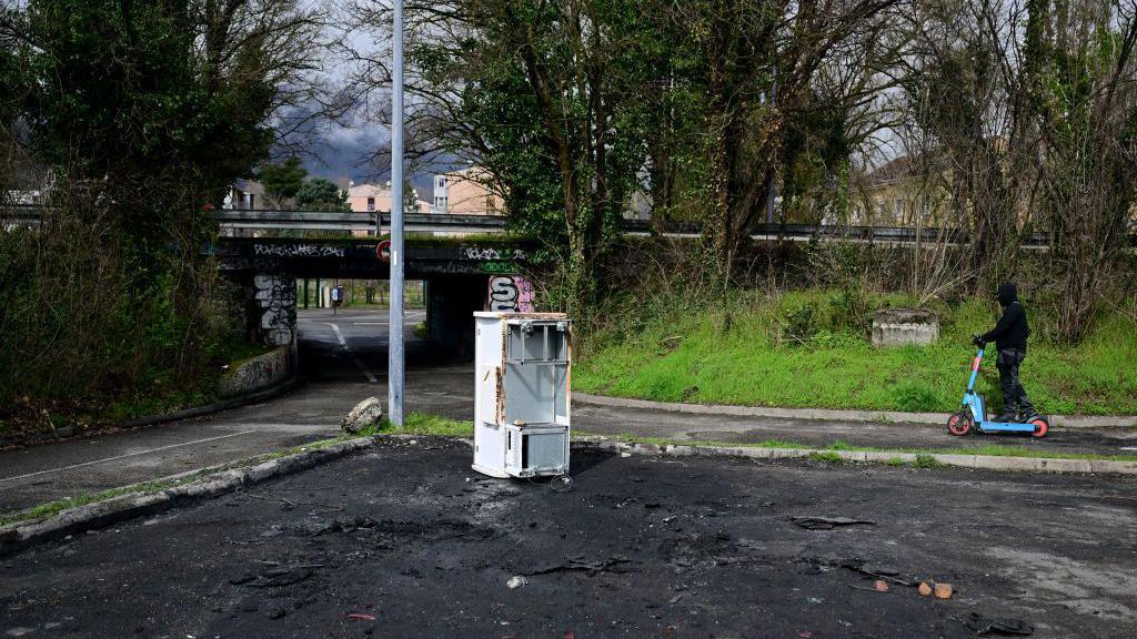 A person rides a scooter by the site where a burnt stolen car was founded linked to the murder of Jean-Pierre Maldera 