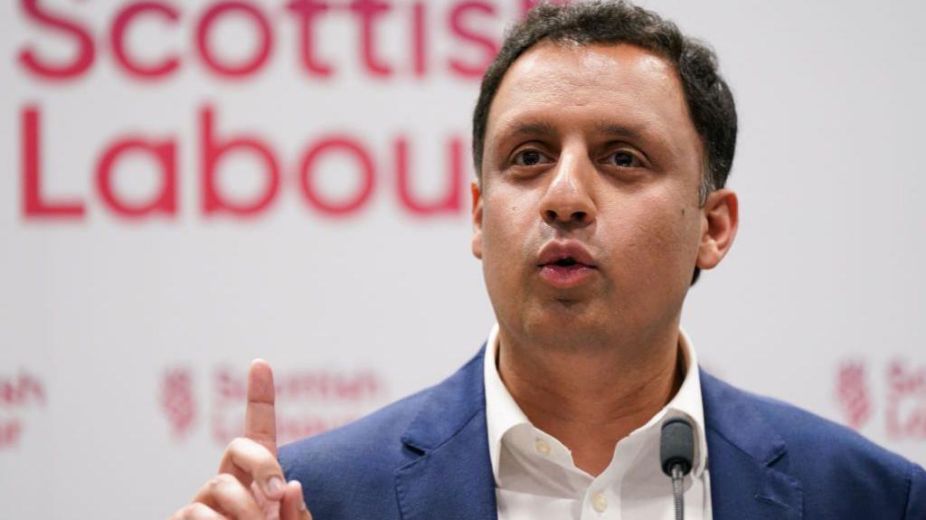 Anas Sarwar raising his finger in front of a red Scottish Labour sign 