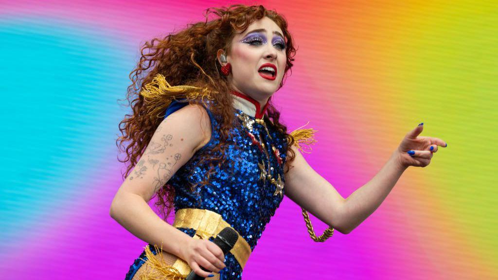 Chappell Roan performing live on stage, holding a microphone in one hand and gesturing to the crowd with the other. She's wearing bright purple eye shadow, red lipstick and a glittery blue outfit with gold trim in front of a technicolour backdrop.