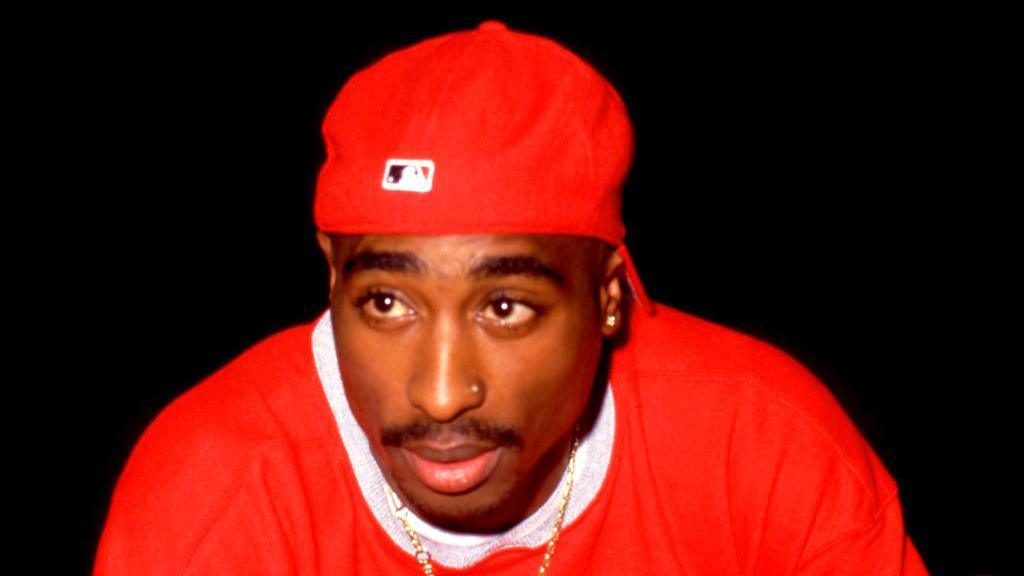Rapper Tupac Shakur, pictured wearing a red sweater and red backwards cap with gold jewellery around his neck