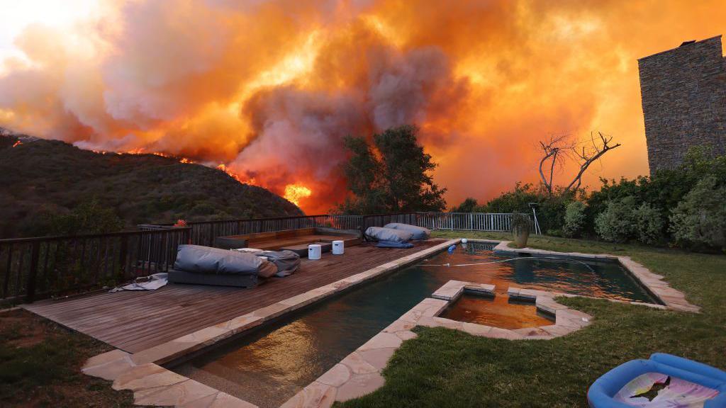 Wildfires in LA. A swimming pool is seen in the foreground as flames surround the forest behind it. 