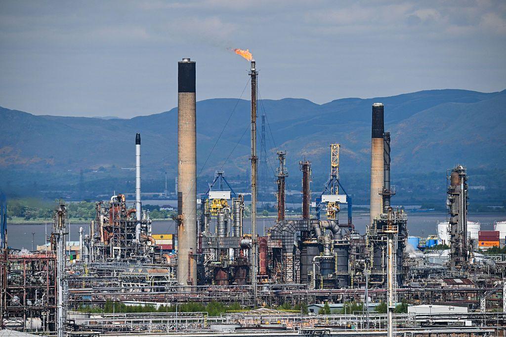 Grangemouth oil refinery stands in front of the Clyde river with mountains in the background. The refinery has several tall towers, with one emitting flames