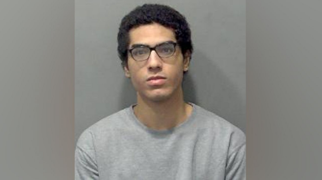 Custody image of Nicholas Prosper. He is wearing a grey sweatshirt and dark-framed glasses.