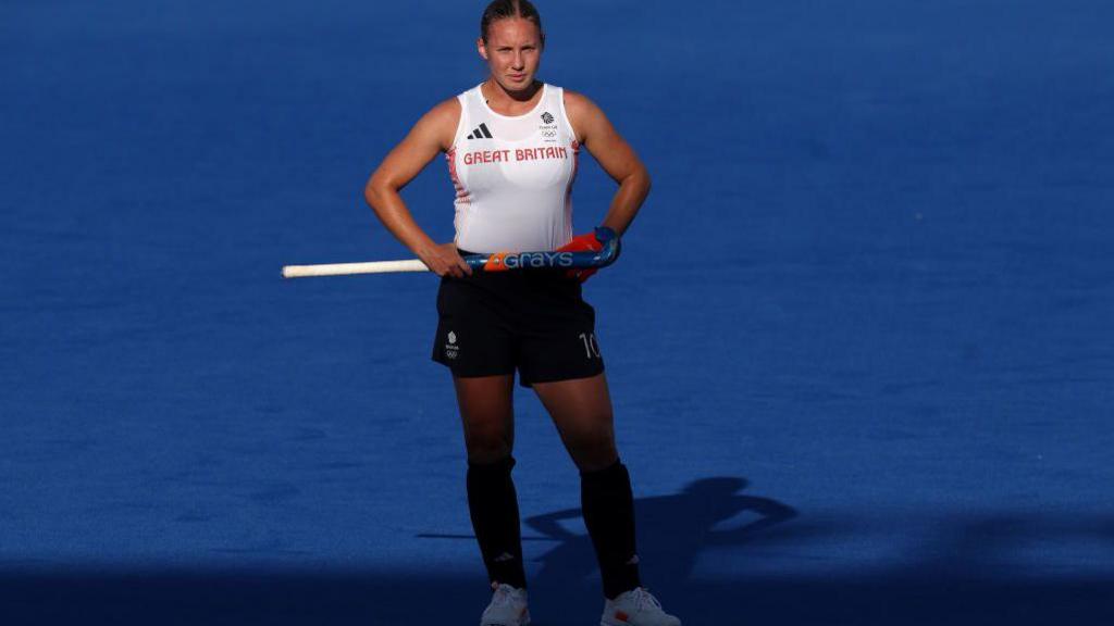 Hockey player Sarah Robertson in Great Britain white top and navy skirt stands with hands on hips, and holding hockey stick, with blue pitch in background.