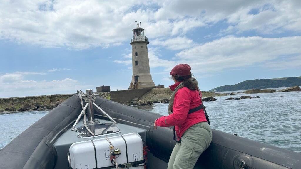 Jennifer Kloester on a small black boat on water looking out towards the lighthouse. She is wearing a red jacket, red cap and green trousers. The engine of the small boat is grey and white. To the right, at the end of the breakwater, is the lighthouse which is grey.