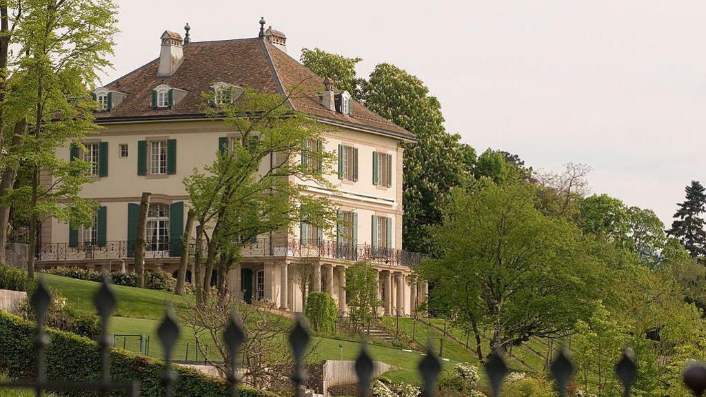 The Villa Diodati is a handsome, large, four-storeyed house with cream plaster and green window shutters, standing in well-tended gardens. A spiky green metal fence can be seen in the foreground.
