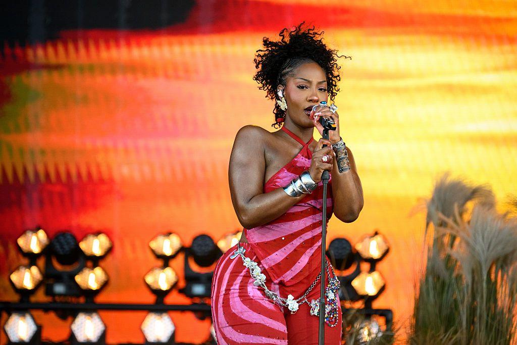 A woman wearing a form-fitting pink and red outfit sings into a microphone on stage. On the set behind her is a sunset-coloured screen, stage lights and some plants.