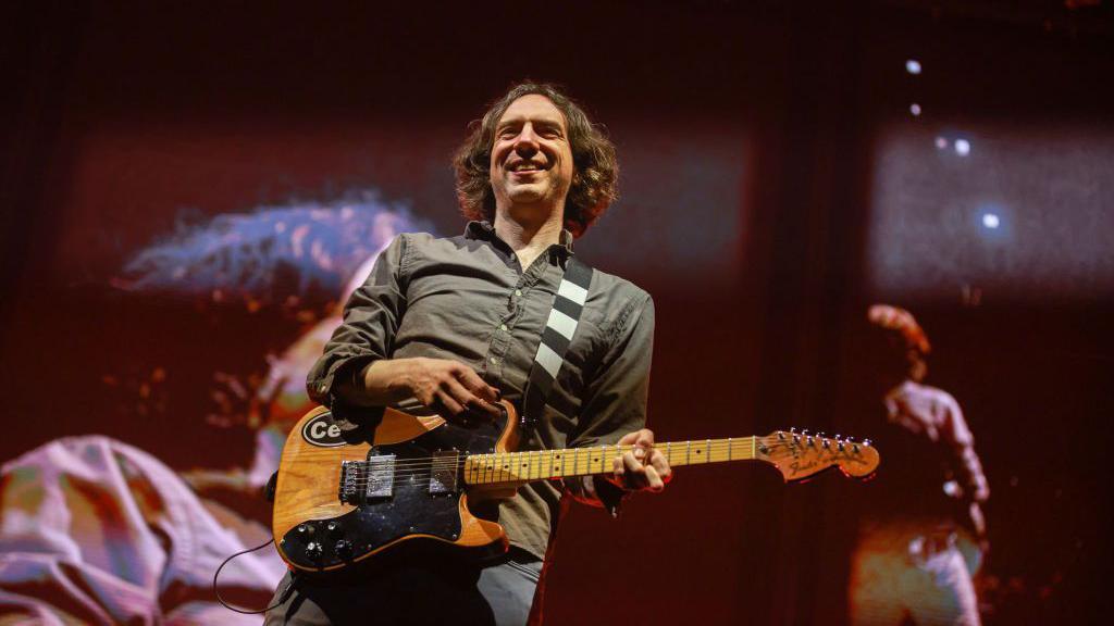 Snow Patrol Performs At 3Arena, Dublin.  Lead singer Gary Lightbody is holding an electric guitar and smiling.  He has wavy, greying dark hair and is wearing a grey shirt and jeans. He also appears on large TV screens at the back of the stage. 