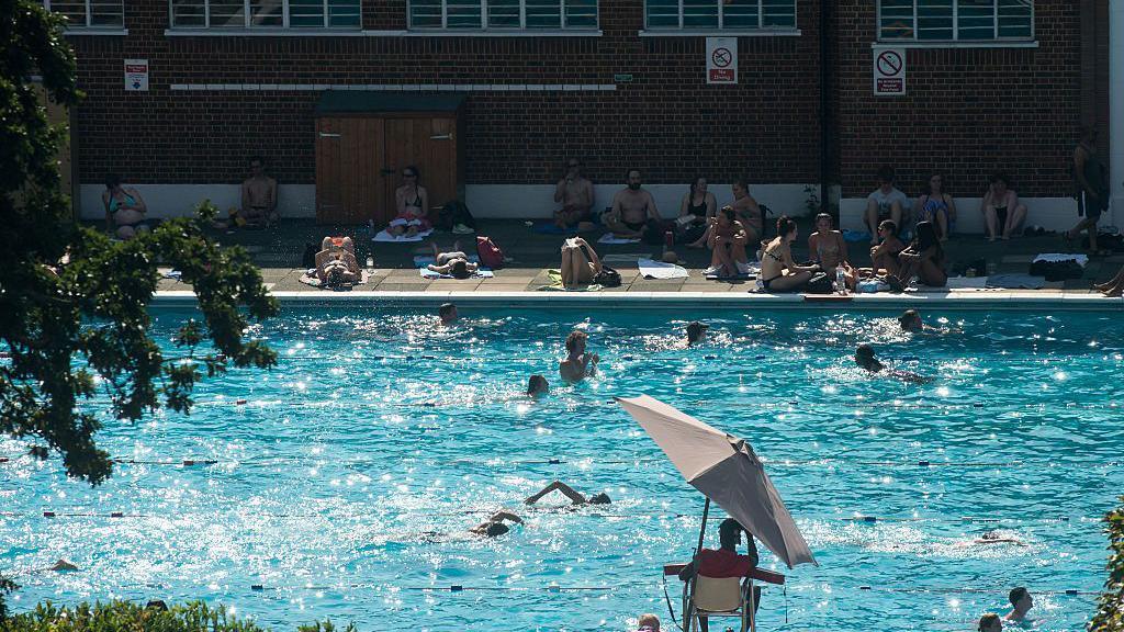 Dozens of people swim and sunbathe around the outdoor pool at Brockwell Lido on a sunny day, with a lifeguard seated under a parasol in the foreground.