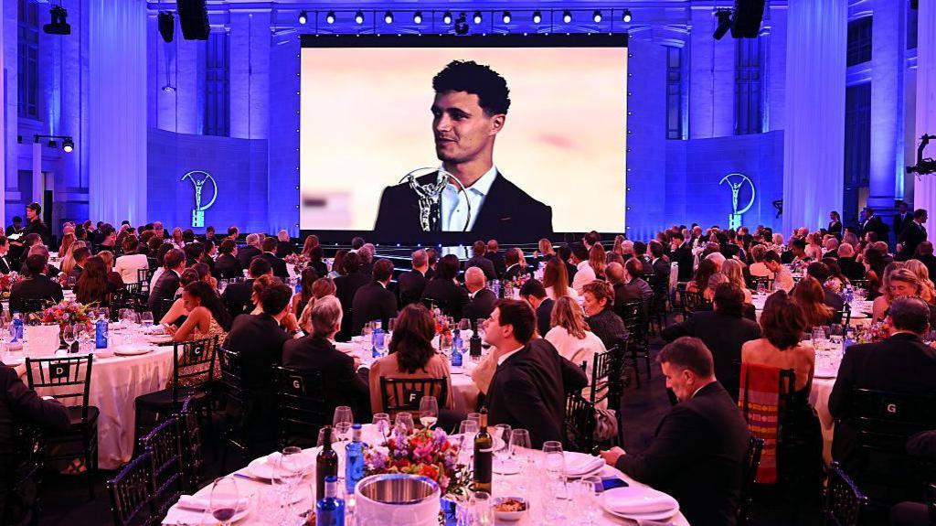 In a large room full of people seated at circular dinner tables decked with wine glasses, flowers and wine bottles, Lando Norris appears on a big screen thaking Laureus for his award.