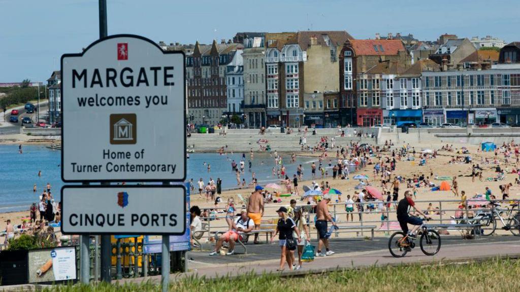 A beach on a sunny day with lots of people out and about. In the foregound there is a sign which reads "welcome to Margate".