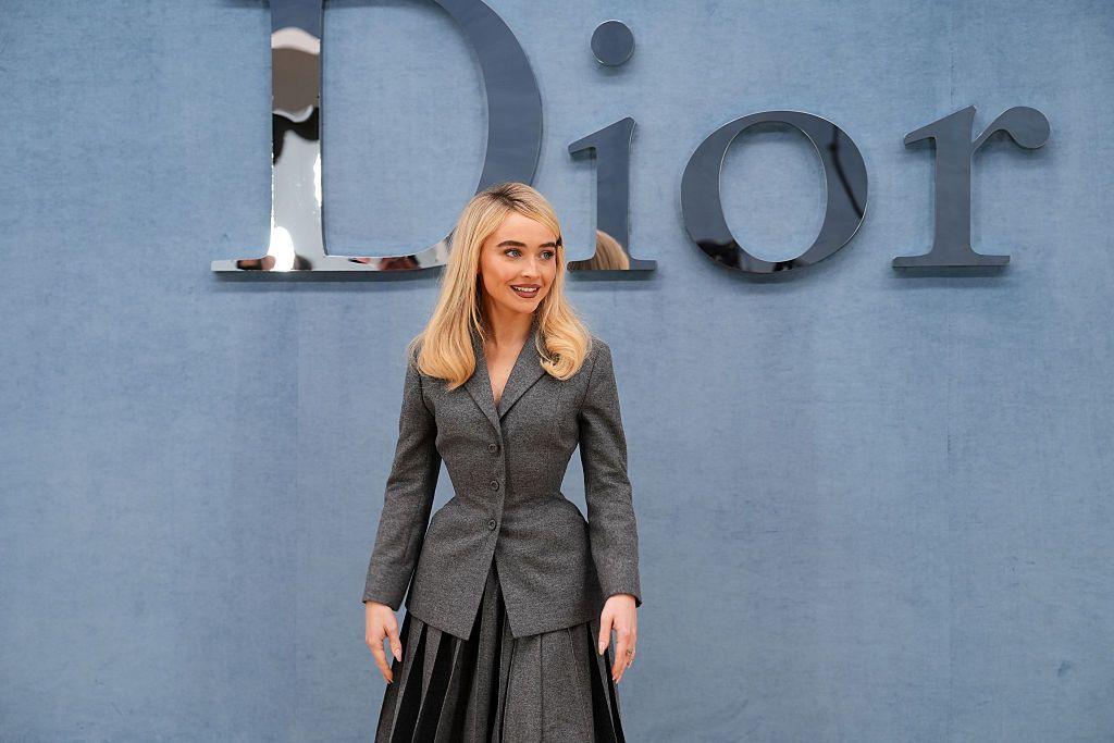 Sabrina Carpenter. A blonde woman wearing a grey suit stands in front of a silver Dior sign.