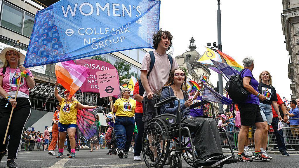 A woman in a wheelchair holding an LGBT+ flag takes part in the parade, surrounded by other participants. 