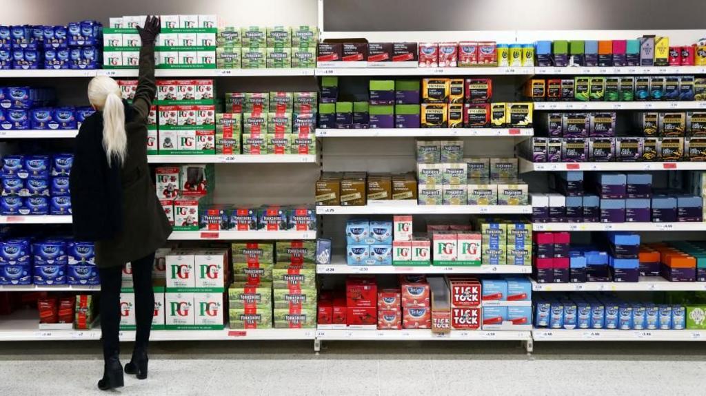 A person stands in a supermarket aisle and reaches for tea