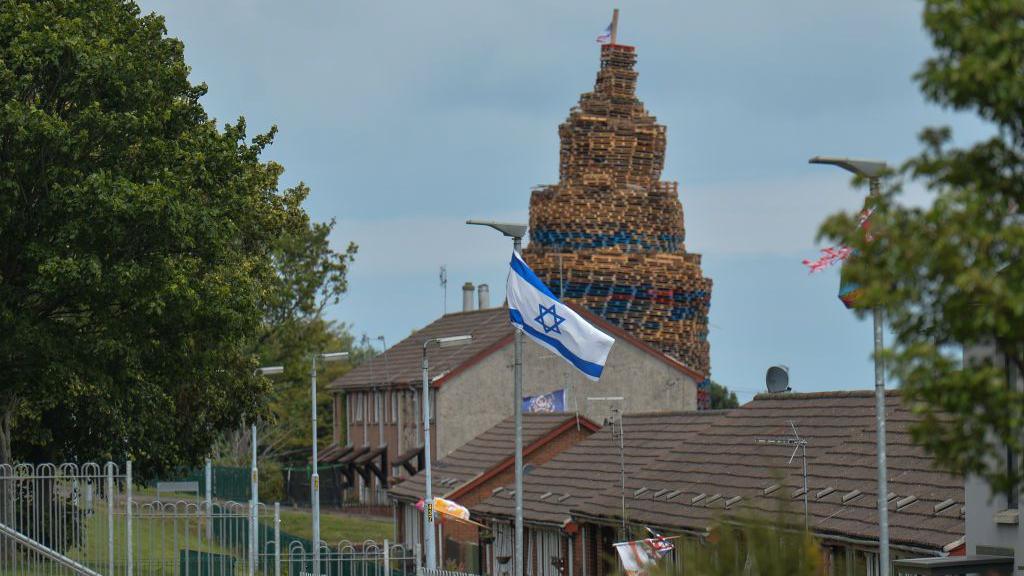 An Israeli flag flying from a lamppost. In the background some houses and behind that a bonfire made of wooden pallets.
