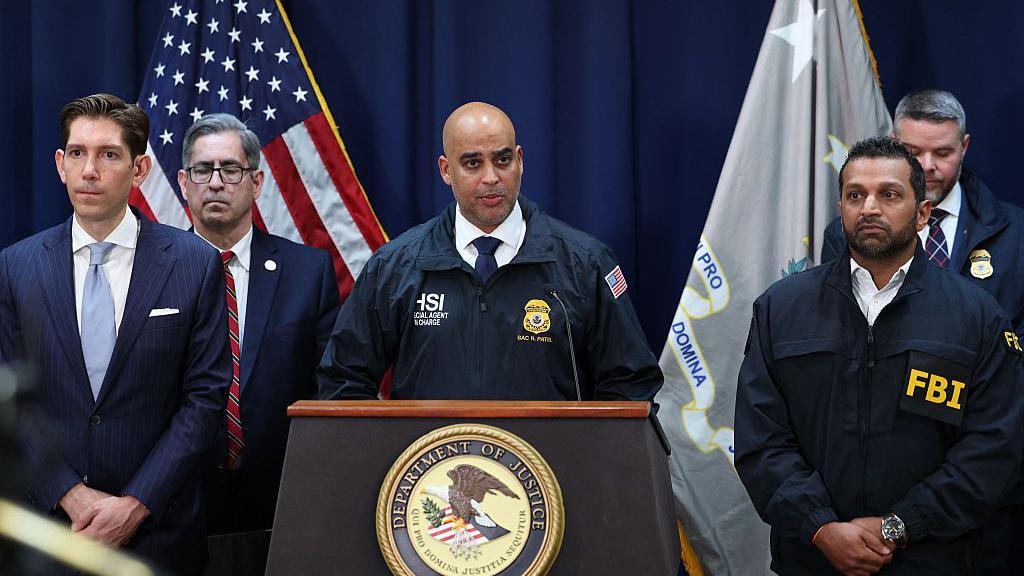 Ricky Patel pictured standing at a podium, with other US officials, including FBI director Kash Patel, standing at each side of him. He is wearing a white shirt, navy tie and jacket with "HSI" branding.