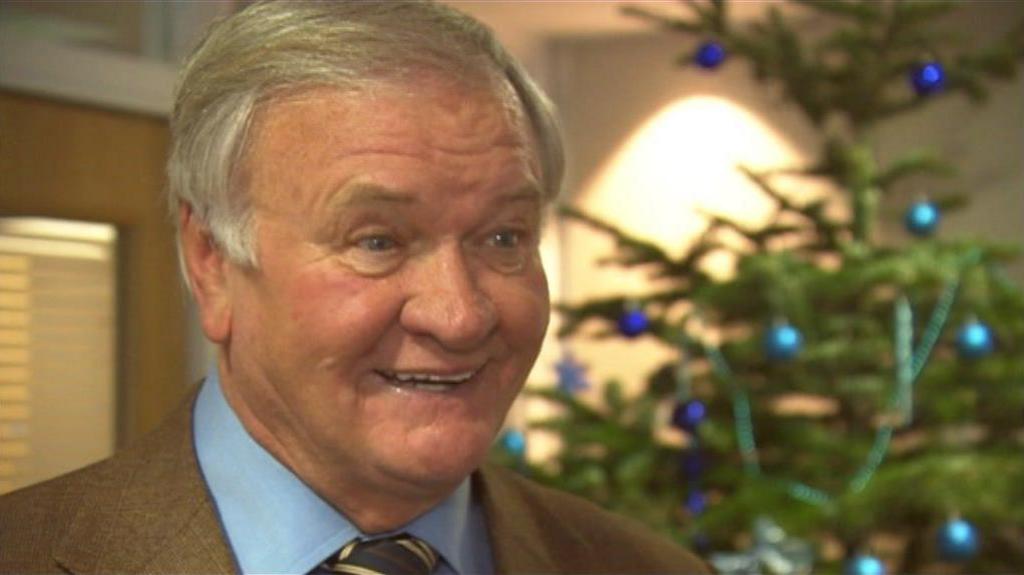 A man wearing a blue shirt and tie stands with a Christmas tree in the background.