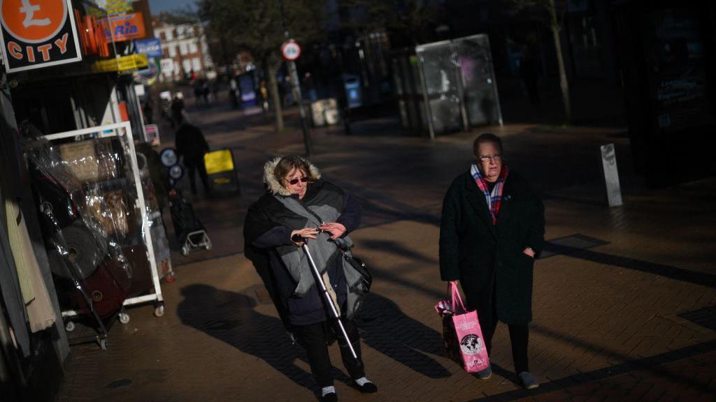 Women walk down the high street in Grays on January 18, 2023. They are wearing black coats and one is holding a walking stick.